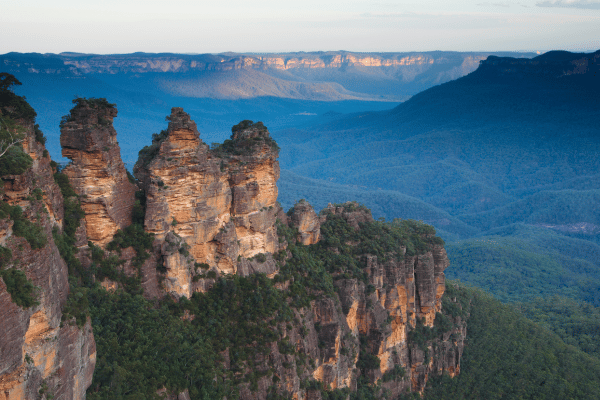 a rocky cliff with trees and mountains in the background with Three Sisters in the background - Blue Mountains, Australia