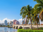 Scenic waterfront view in West Palm Beach, Florida, featuring palm trees, a bridge, and modern buildings under a bright blue sky.
