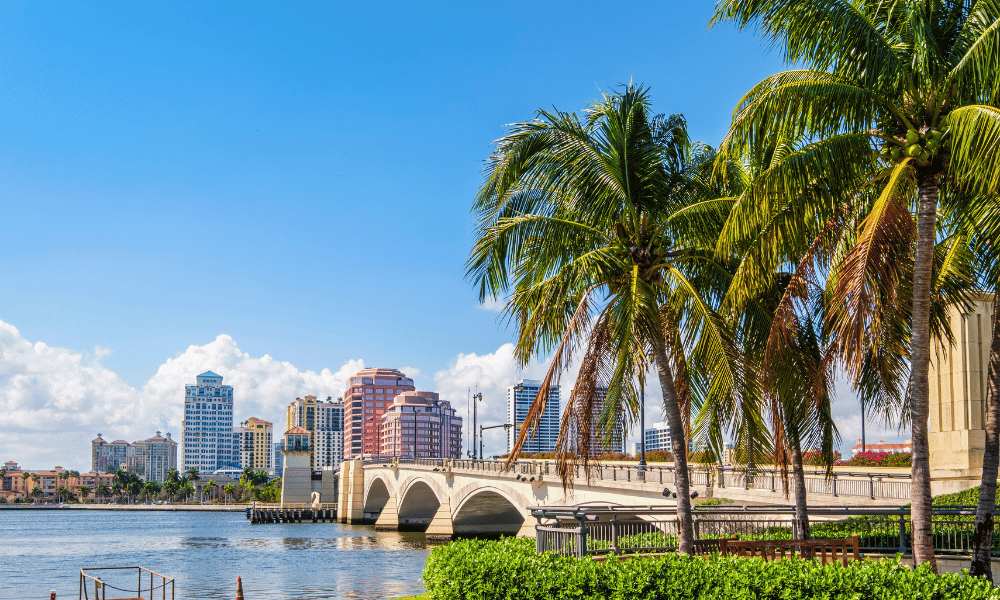 Scenic waterfront view in West Palm Beach, Florida, featuring palm trees, a bridge, and modern buildings under a bright blue sky.